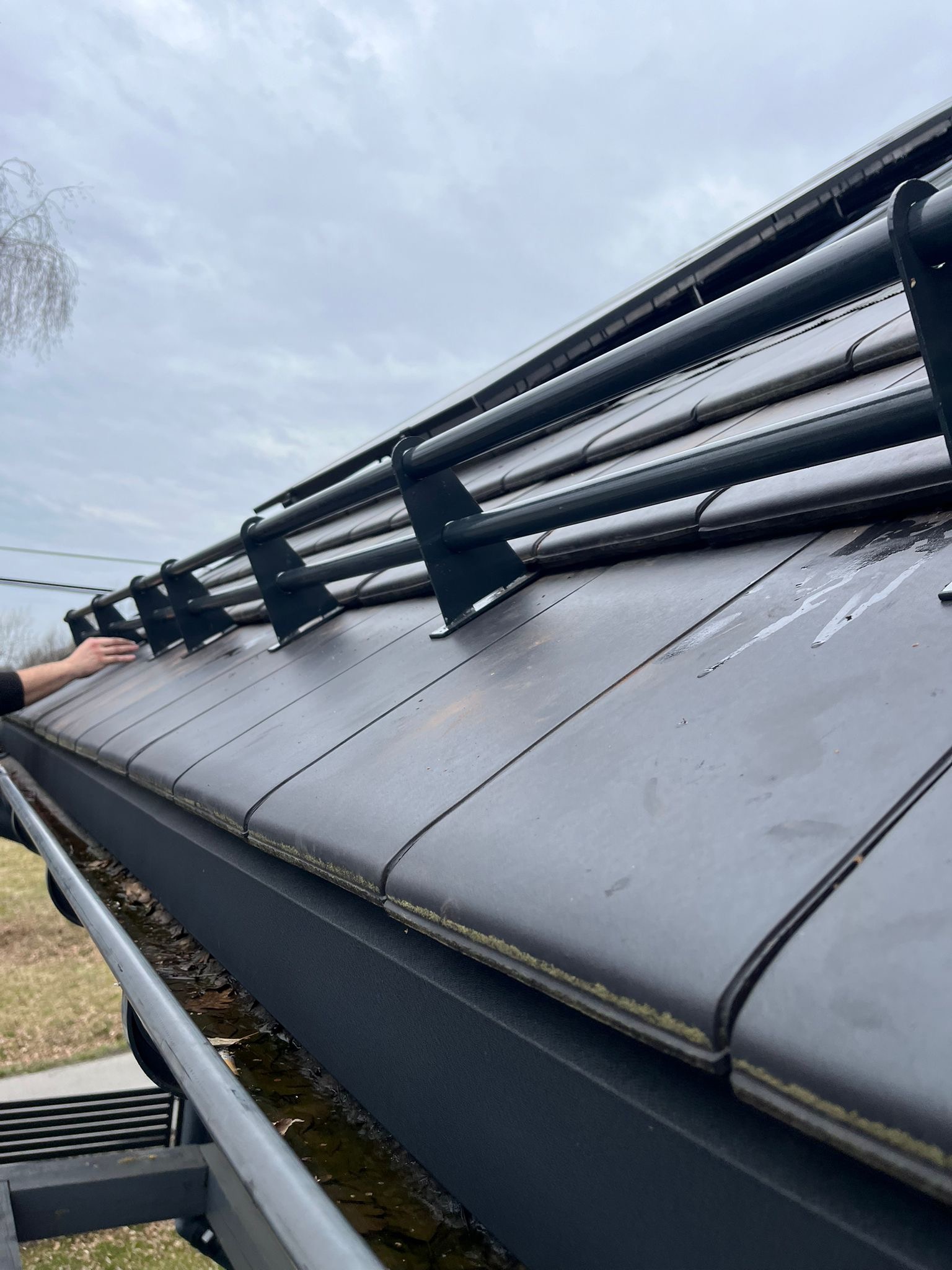 Czyszczenie rynny dachowej z nagromadzonych liści, widoczne metalowe zabezpieczenie przed śniegiem, dachówka ceramiczna i fragment dłoni pracownika.