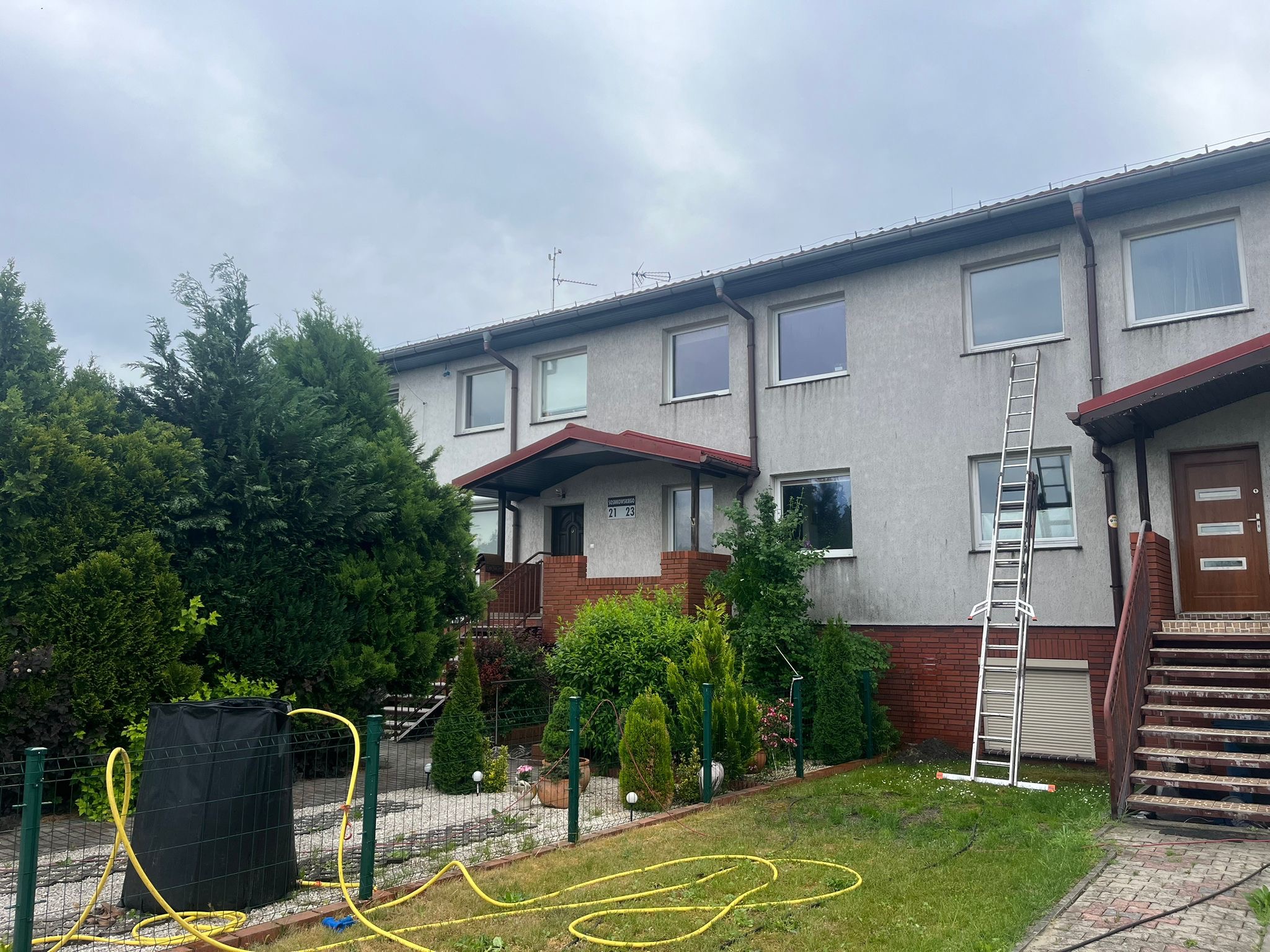 Dwupiętrowy budynek z szarą elewacją, widoczną drabina oparta o ścianę, żółty wąż ogrodowy leżący na trawniku, oraz zbiornik na deszczówkę w ogrodzie.