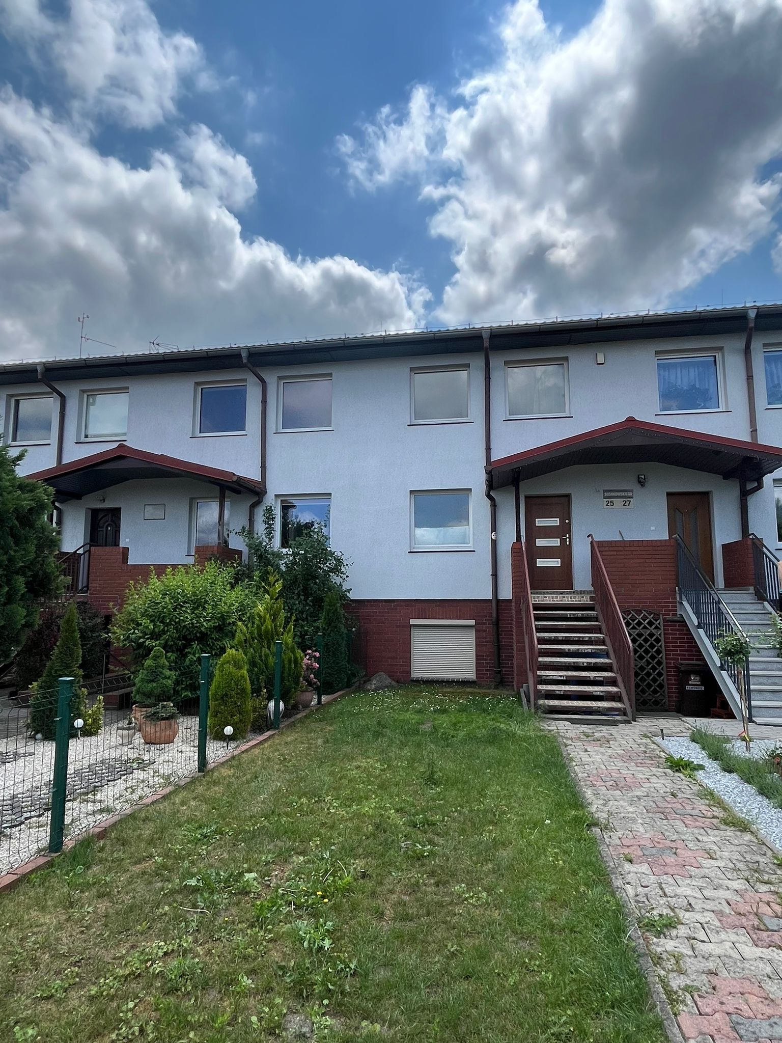 Szereg domów z szarą elewacją, brązowymi rynnami i czerwonymi ceglanymi elementami, widok z poziomu trawnika w pochmurny dzień.