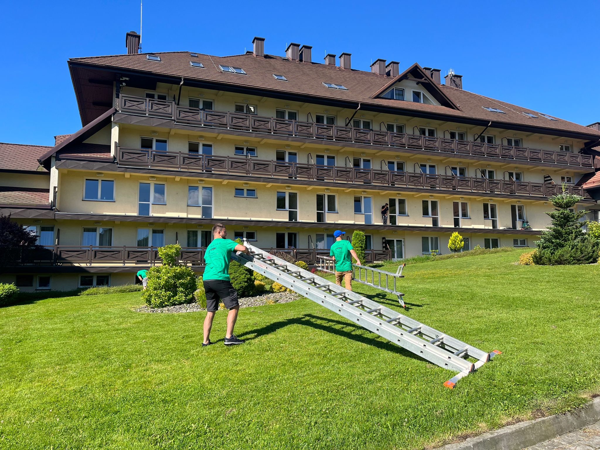 Dwóch mężczyzn w zielonych koszulkach transportuje rozłożoną drabinę na trawniku przed trzypiętrowym budynkiem z brązowym dachem i balkonami.