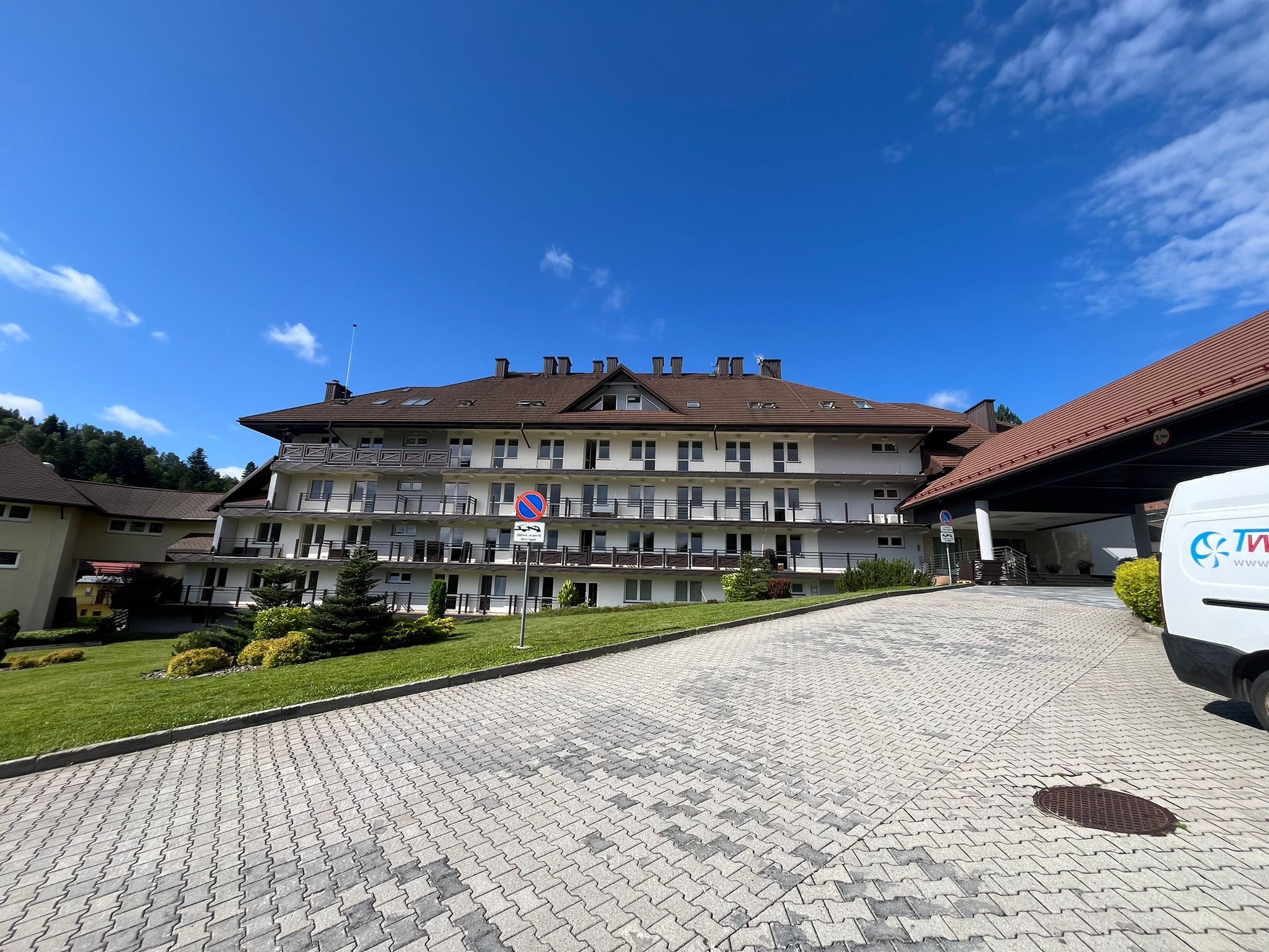 Trzypiętrowy budynek z balkonami i brązowym dachem, widok z dołu, w tle błękitne niebo, na pierwszym planie podjazd z kostki brukowej i fragment zaparkowanego samochodu dostawczego.