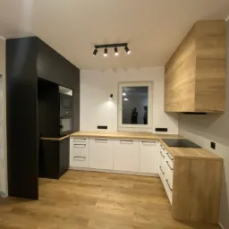 Nowoczesna kuchnia z zabudową w kolorach czerni, bieli i drewna, oświetlona reflektorami, z drewnianą podłogą i widokiem przez okno.