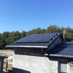 Panele słoneczne na dachu domu z blachy trapezowej, antena telewizyjna, w tle las iglasty pod błękitnym niebem.