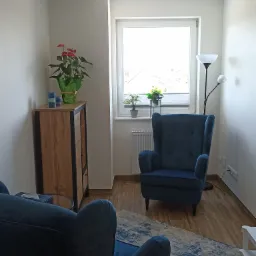 Jasny pokój z dwoma granatowymi fotelami typu uszak, drewnianą komodą z kwiatem anturium i lampą podłogową, dywanem z wzorem w odcieniach szarości i błękitu oraz roślinami w doniczkach na parapecie.