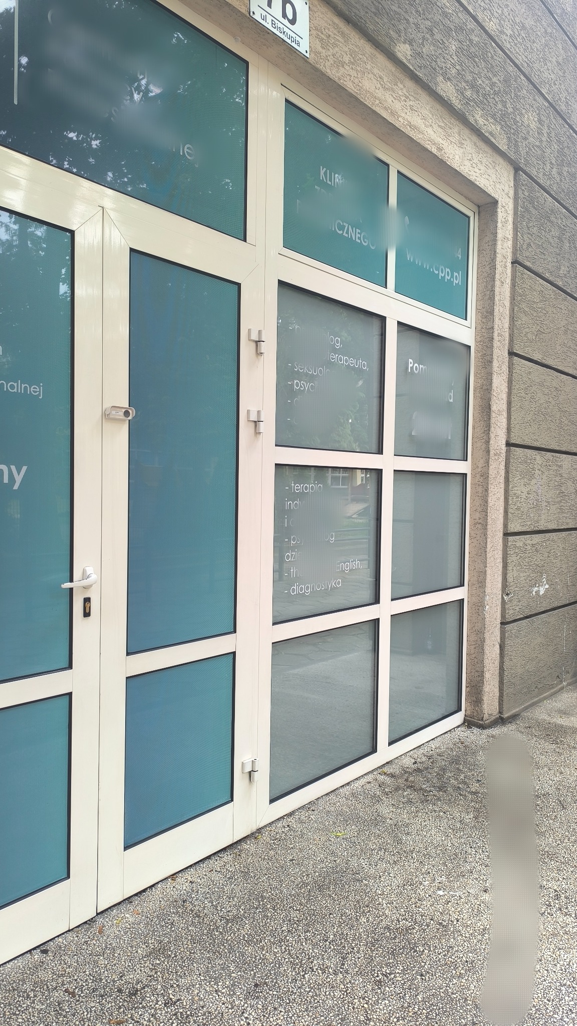 Wejście do kliniki psychologicznej z białymi ramami okiennymi i niebieskimi szybami z informacjami o usługach: terapia, psycholog, seksuolog, diagnostyka.