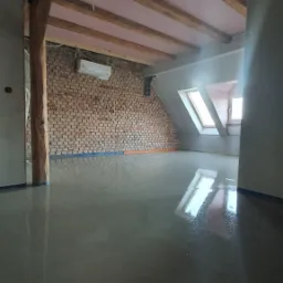 Wnętrze poddasza z surową ceglaną ścianą, drewnianymi belkami stropowymi z izolacją, nową wylewką podłogową i oknami dachowymi, widoczny klimatyzator.