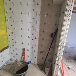 Ściana łazienki w trakcie układania płytek ceramicznych, widoczne krzyżyki dystansowe, poziomica, wiadro z zaprawą i narzędzia glazurnicze na podłodze, częściowo ułożony wzór dekoracyjny.