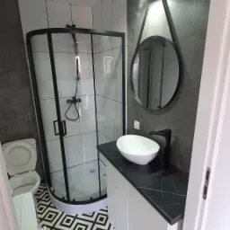 Nowoczesna łazienka z kabiną prysznicową z czarnymi ramami, umywalką nablatową na szafce z blatem imitującym marmur, okrągłym lustrem z czarną obwódką i wzorzystą czarno-białą podłogą.