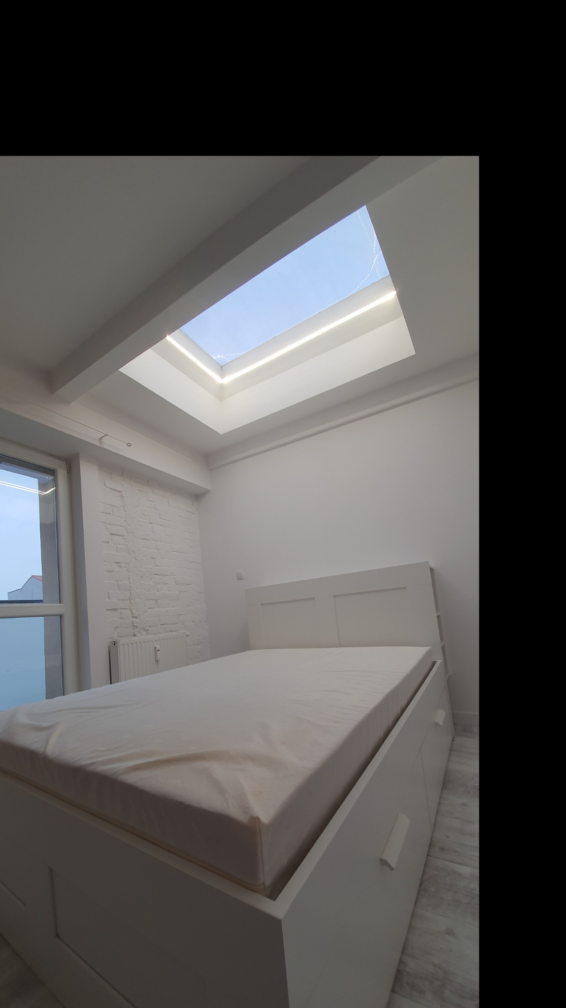 Jasne wnętrze sypialni z białymi ścianami, ceglaną ścianą, dużym oknem i łóżkiem z szufladami, oświetlone światłem wpadającym przez okno dachowe z listwą LED.