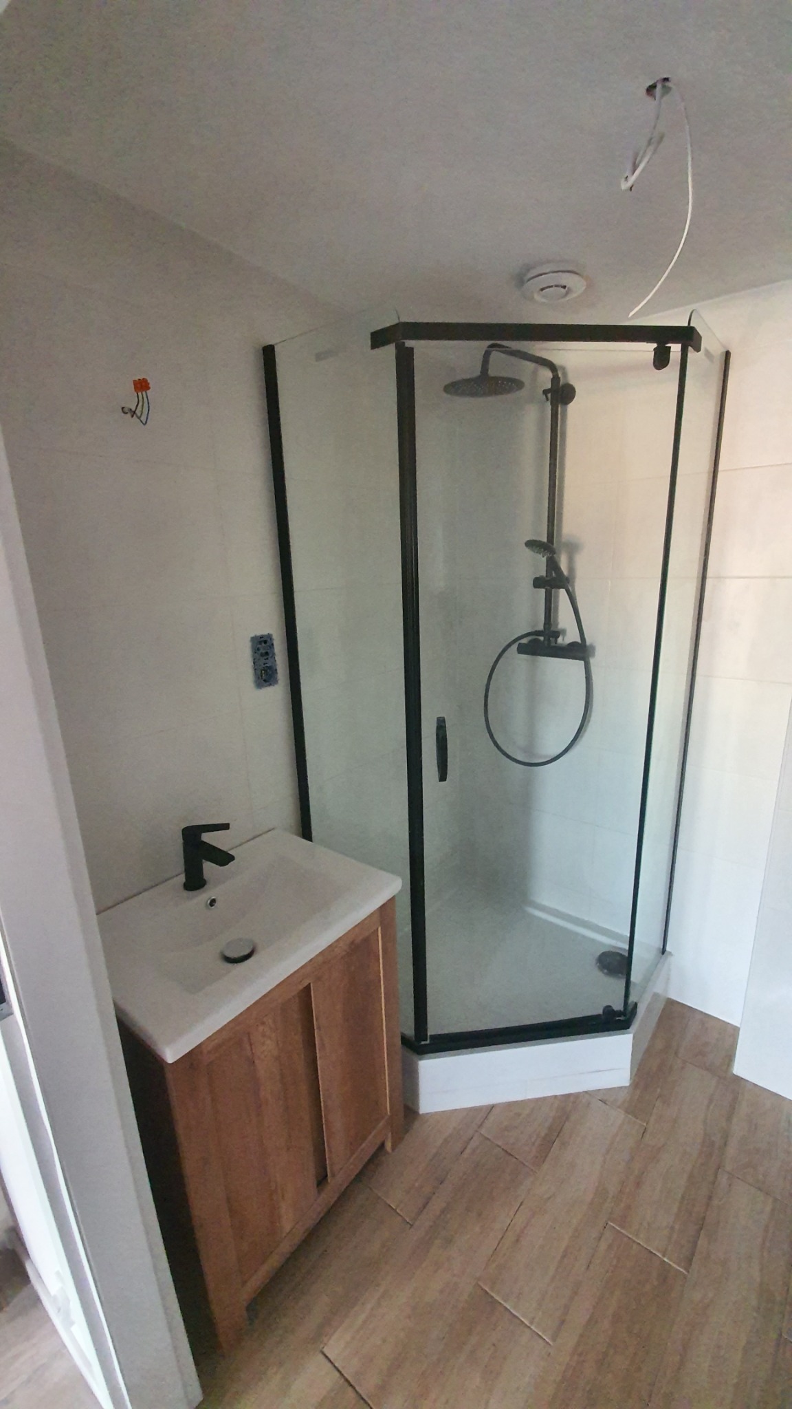 Nowoczesna kabina prysznicowa z czarnymi profilami i deszczownicą, umywalka z szafką z naturalnego drewna, jasne płytki na ścianach i podłodze ułożone w jodełkę, widoczne niedokończone prace...