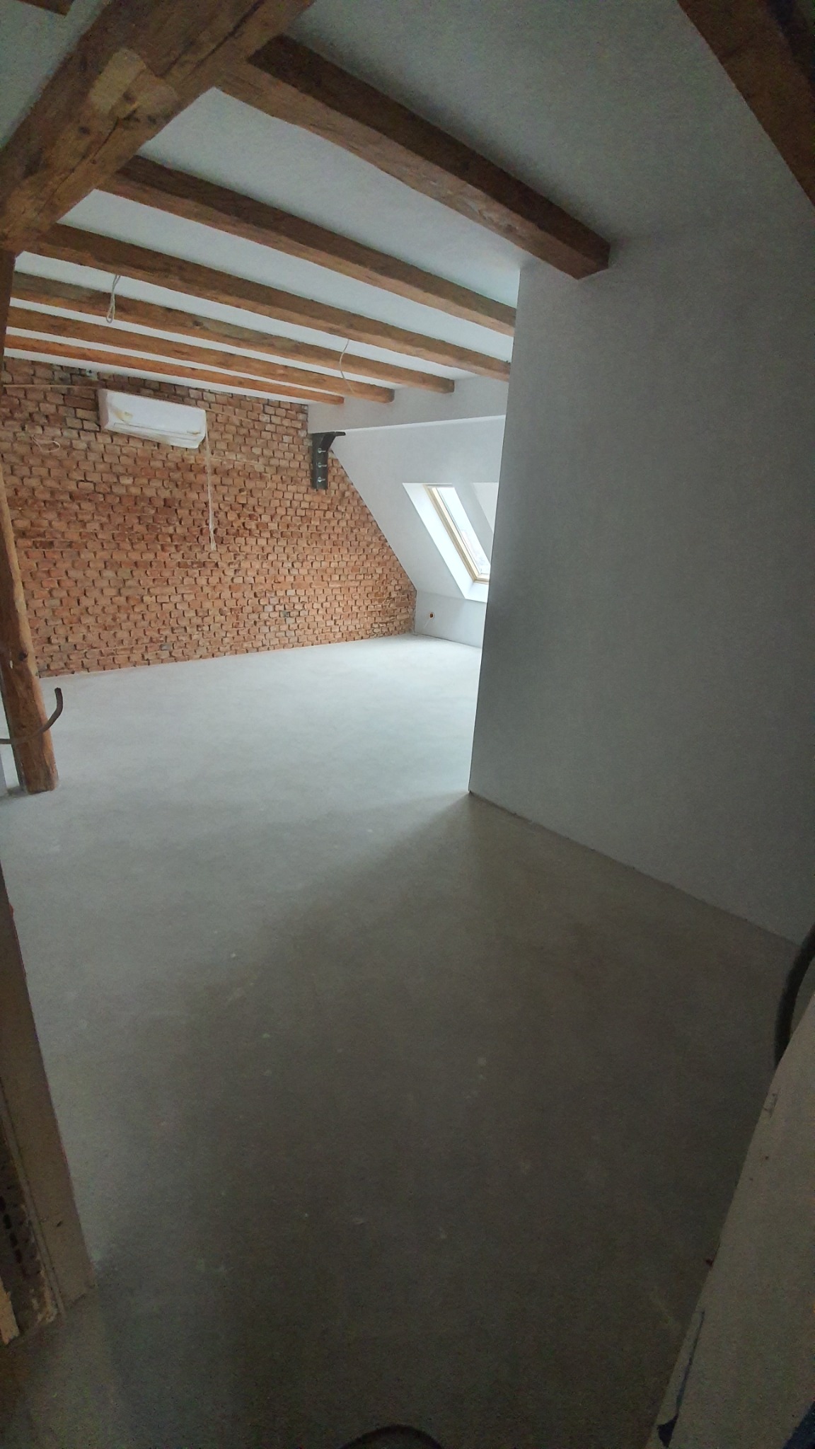 Poddasze po remoncie z odsłoniętą ścianą z czerwonej cegły, drewnianymi belkami stropowymi, gładzią na ścianach i oknem dachowym.