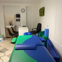 Wnętrze gabinetu z zielono-niebieskim stołem do masażu, krzesłami, umywalką i dekoracją z mchu na ścianie.