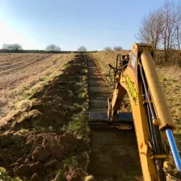 Żółta koparka gąsienicowa marki CAT podczas wykonywania wykopu w długim, wąskim rowie na polu, widok z kabiny operatora na linię horyzontu pod błękitnym niebem.