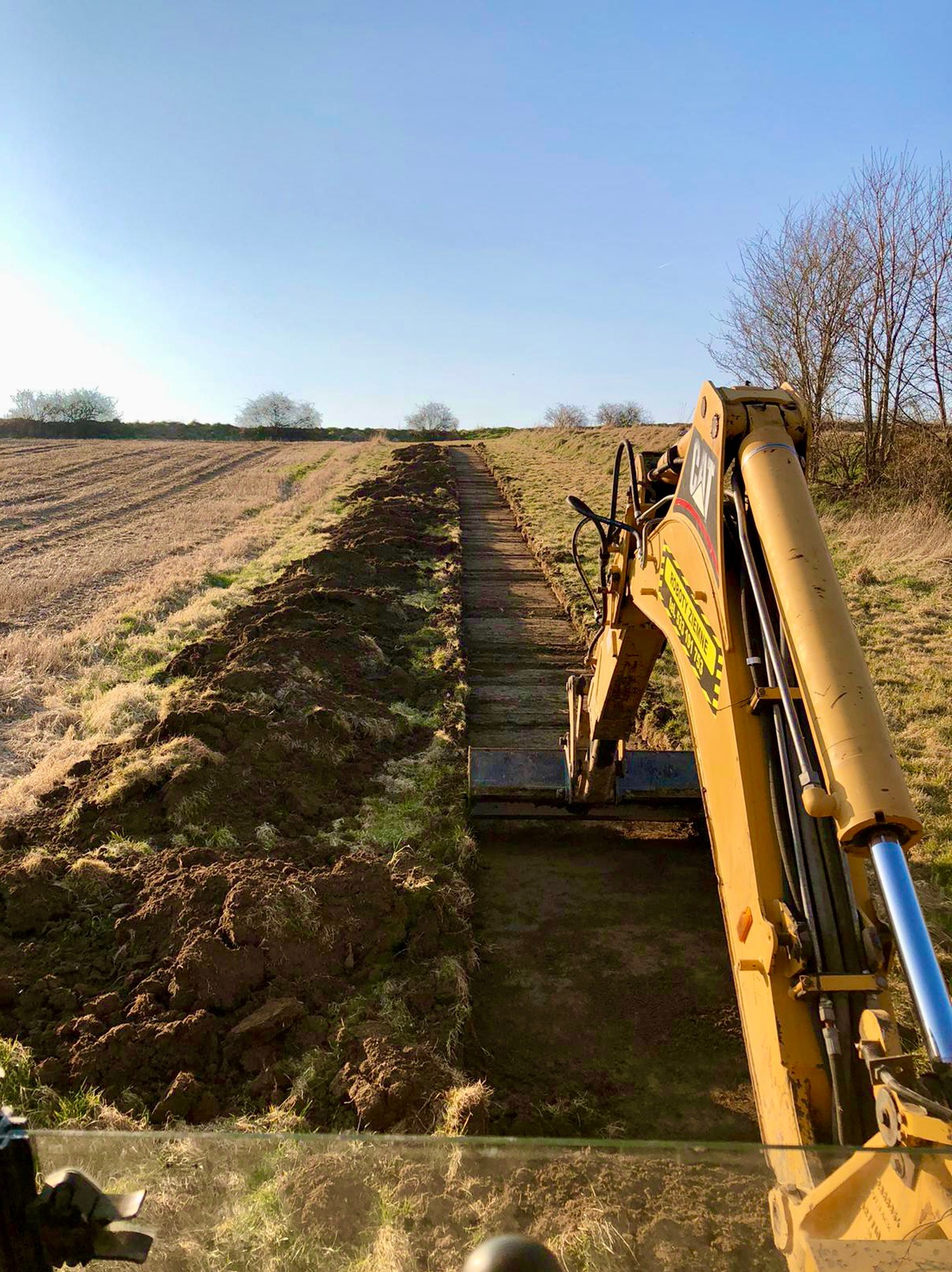 Żółta koparka gąsienicowa marki CAT podczas wykonywania wykopu w długim, wąskim rowie na polu, widok z kabiny operatora na linię horyzontu pod błękitnym niebem.