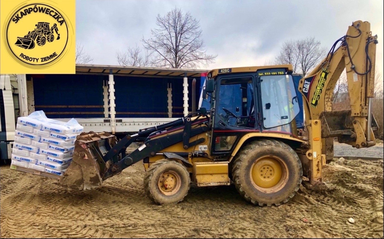 Żółta koparko-ładowarka CAT 428C z podniesioną łyżką, załadowana paletą z workami materiałów budowlanych, obok zaparkowana ciężarówka z niebieską plandeką, na błotnistym terenie.