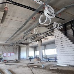 EPI - Wnętrze hali w trakcie budowy z widocznymi trasami kablowymi pod sufitem, zwisającymi rolkami kabli, instalacjami elektrycznymi i fragmentem ściany z białych bloczków.