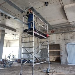 EPI - Elektryk na rusztowaniu montuje koryta kablowe pod sufitem w surowym wnętrzu budynku w trakcie budowy.