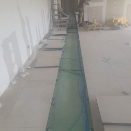 EPI - Montaż instalacji elektrycznej w podłodze technicznej budynku w trakcie budowy, widoczne kable w korycie, płyty podłogowe, narzędzia i pracownik w tle.