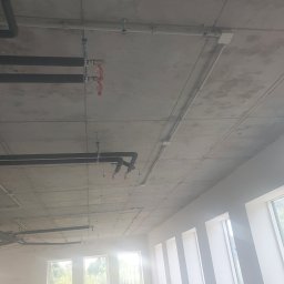 EPI - Betonowy sufit z widocznymi czarnymi rurami instalacyjnymi, białymi rurami elektrycznymi i mocowaniami, w tle rząd okien z widokiem na zieleń.