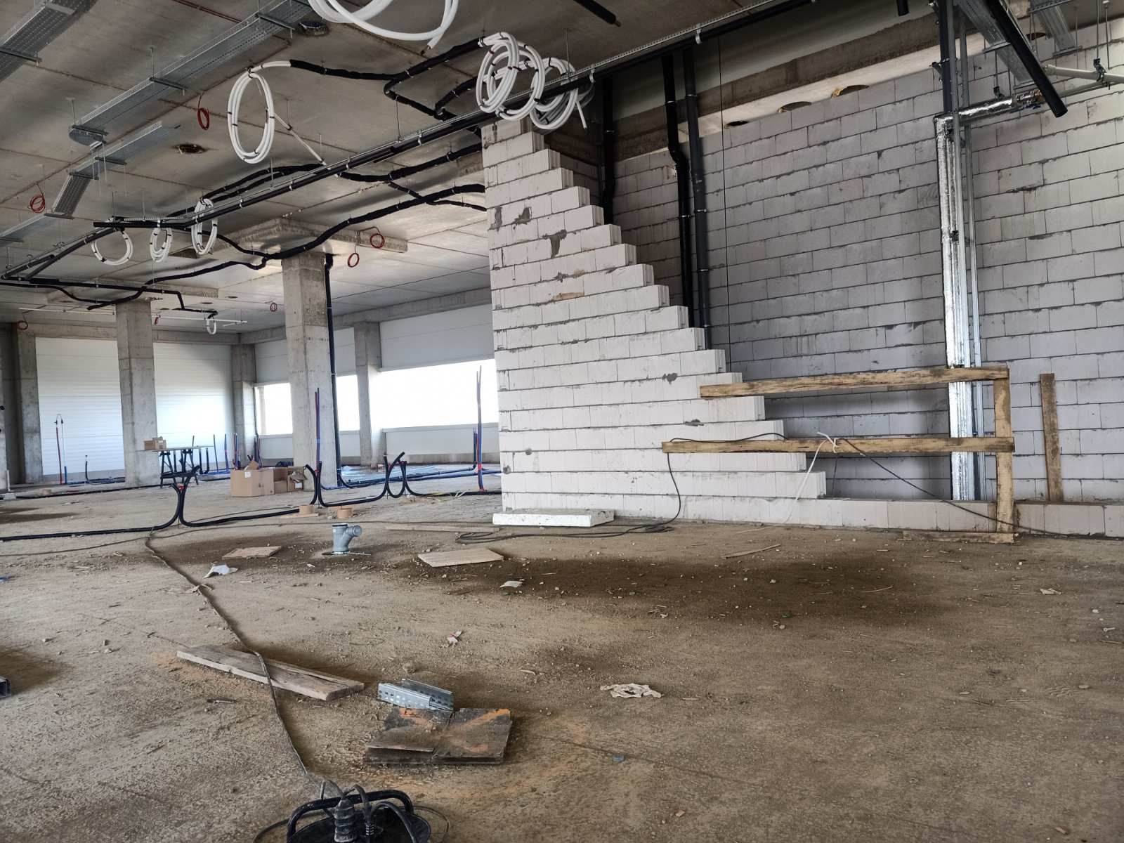 Wnętrze w stanie surowym z widocznymi instalacjami elektrycznymi pod sufitem, koryta kablowe, peszle i przewody, ściana z bloczków betonowych, szalunek schodów, podłoże betonowe.