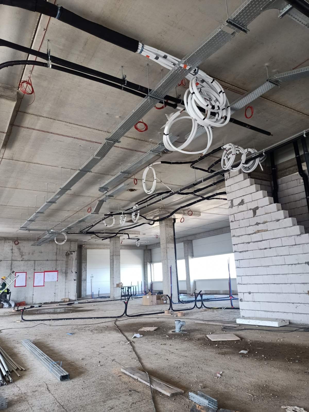 Wnętrze hali w trakcie budowy z widocznymi trasami kablowymi pod sufitem, zwisającymi rolkami kabli, instalacjami elektrycznymi i fragmentem ściany z białych bloczków.
