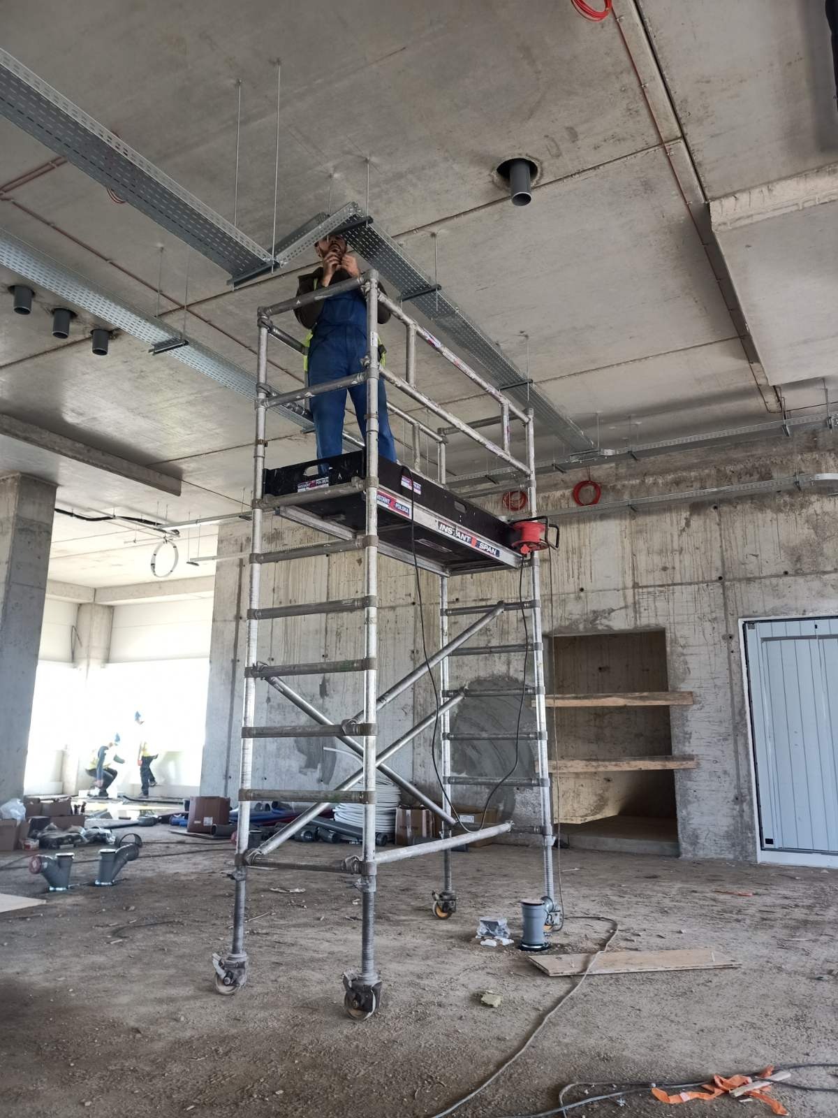 Elektryk na rusztowaniu montuje koryta kablowe pod sufitem w surowym wnętrzu budynku w trakcie budowy.