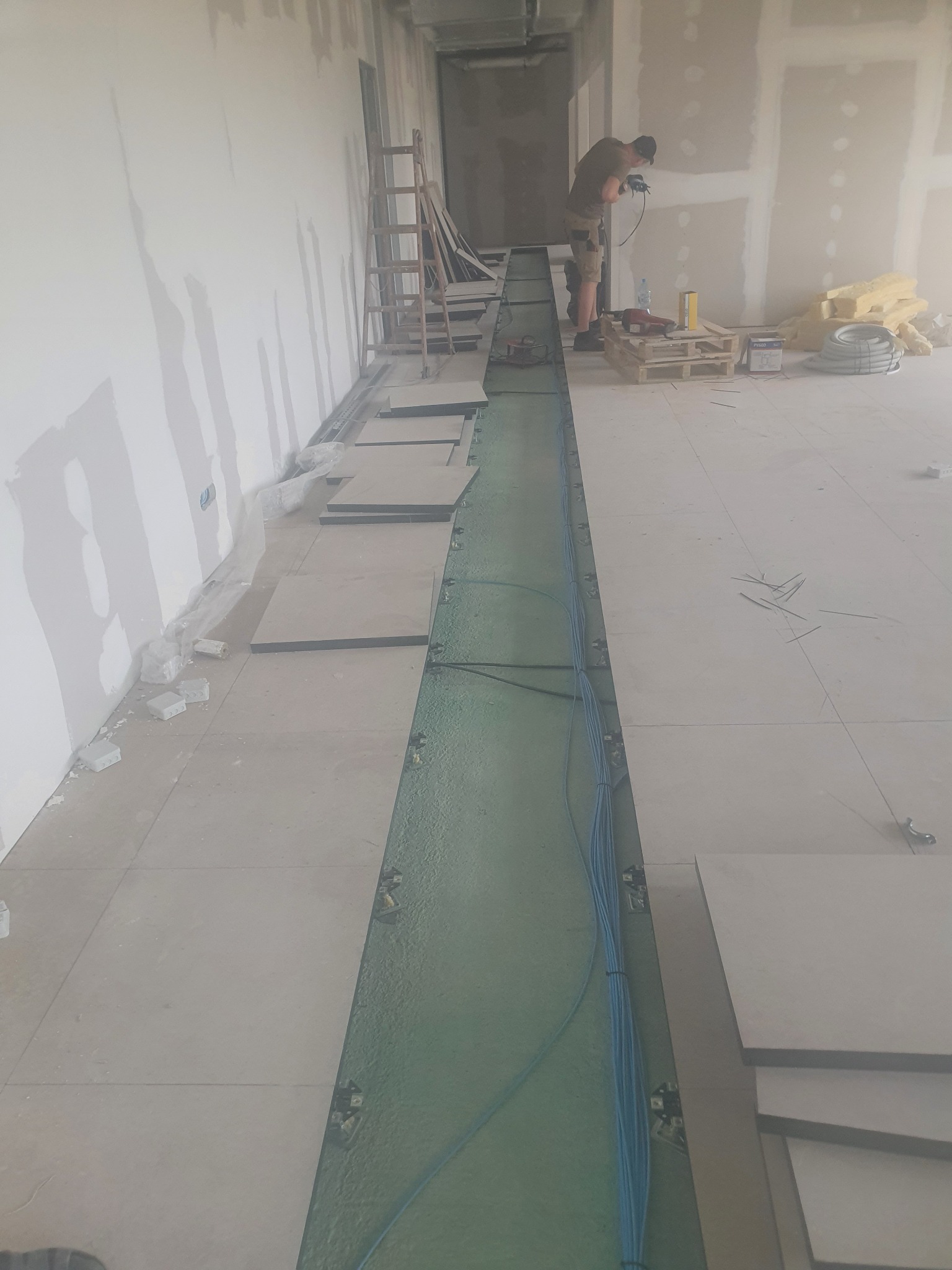 Montaż instalacji elektrycznej w podłodze technicznej budynku w trakcie budowy, widoczne kable w korycie, płyty podłogowe, narzędzia i pracownik w tle.