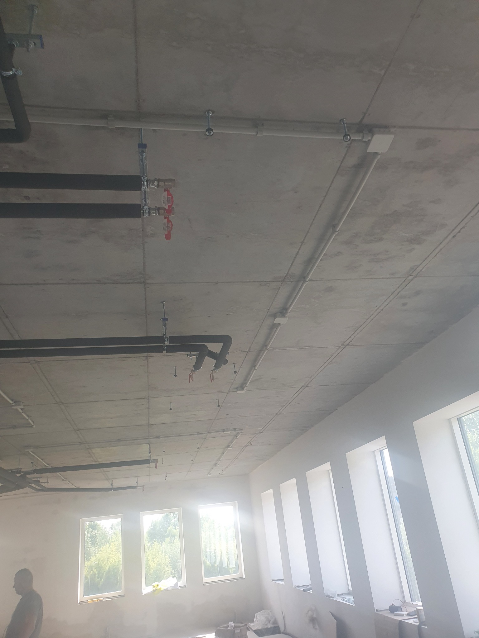 Betonowy sufit z widocznymi czarnymi rurami instalacyjnymi, białymi rurami elektrycznymi i mocowaniami, w tle rząd okien z widokiem na zieleń.