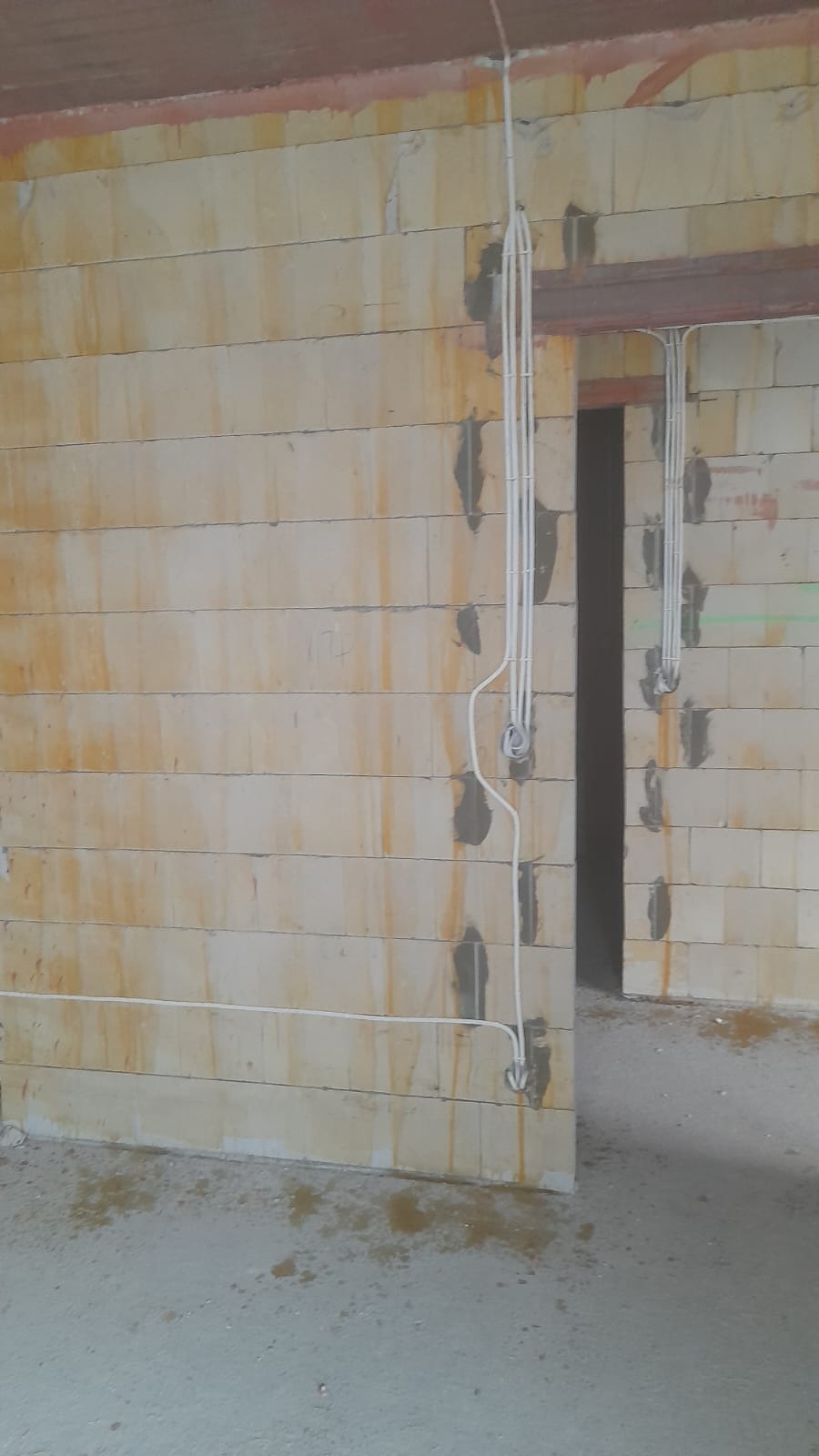 Surowe wnętrze w trakcie prac: widoczne kable elektryczne prowadzone po ścianie z betonu komórkowego, mocowane gipsem, oraz otwór drzwiowy z nadprożem.
