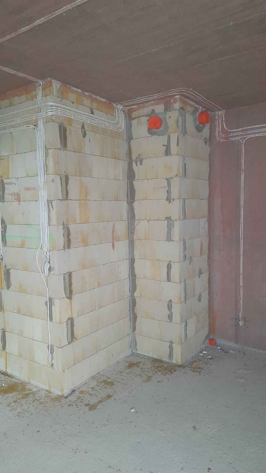 Surowa ściana z betonu komórkowego z widocznymi przewodami elektrycznymi prowadzonymi po powierzchni, pomarańczowe puszki instalacyjne pod sufitem, stan surowy budynku.
