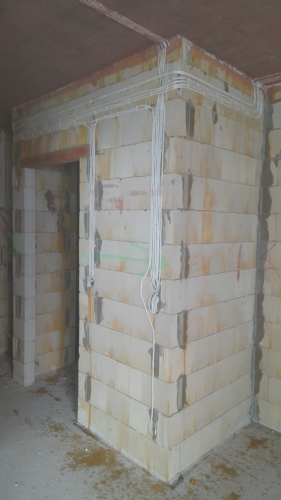 Wewnętrzne ściany budynku w trakcie budowy z widocznymi, równolegle ułożonymi przewodami elektrycznymi pod sufitem oraz pionowymi wiązkami kabli wzdłuż narożnika ściany.