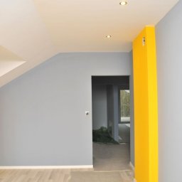 Tomasz mal - Wnętrze po remoncie: szare ściany kontrastują z intensywnie żółtymi akcentami przy oknach i narożnikach, podłoga wyłożona jasnymi panelami, widoczne punkty świetlne w suficie.