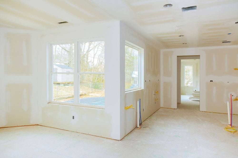 Wnętrze w trakcie remontu: ściany i sufit pokryte szpachlą, widoczne instalacje elektryczne i hydrauliczne, okna z widokiem na ogród.