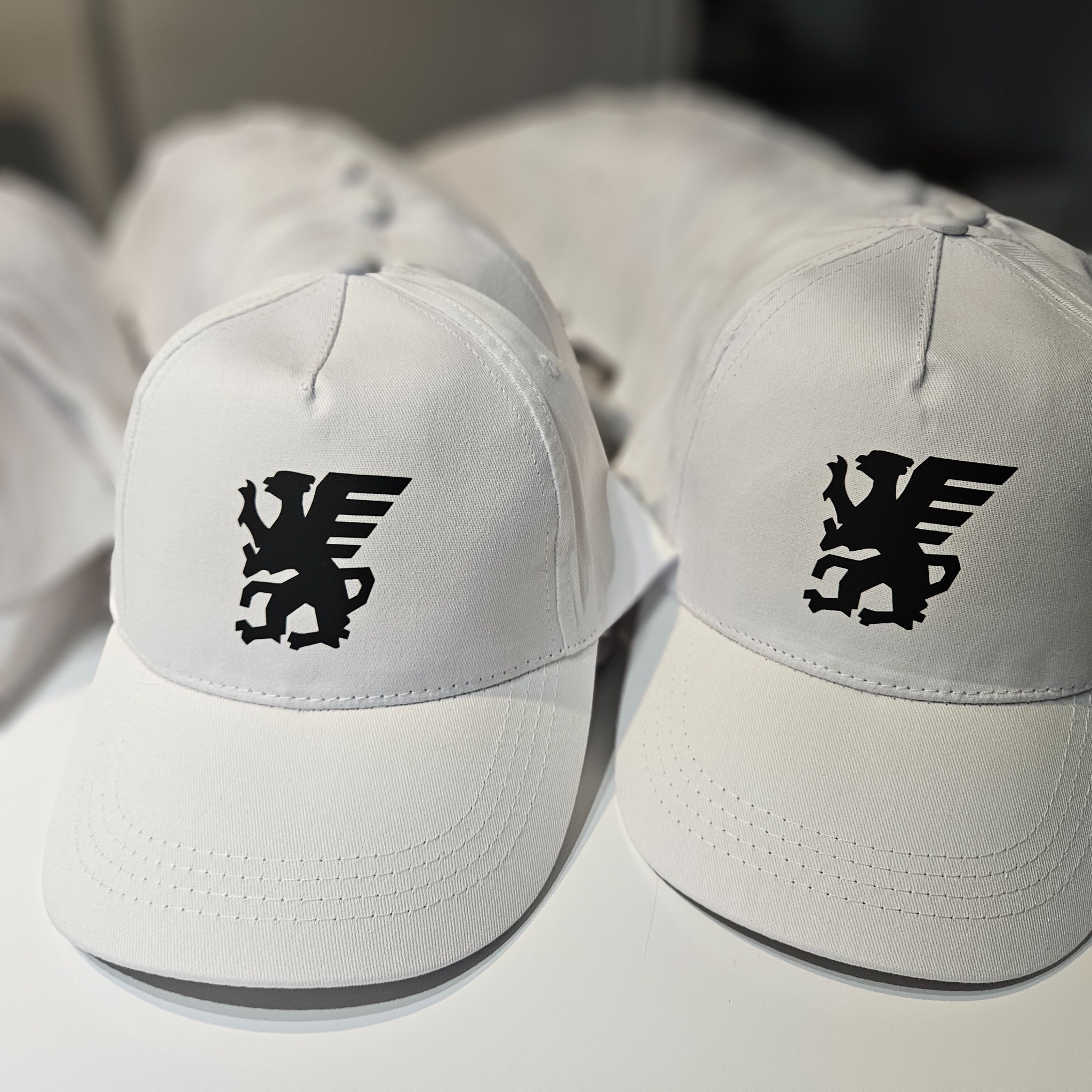 Białe czapki z daszkiem z czarnym nadrukiem gryfa, ułożone rzędami na jasnym tle. Widoczne detale szwów i materiału.