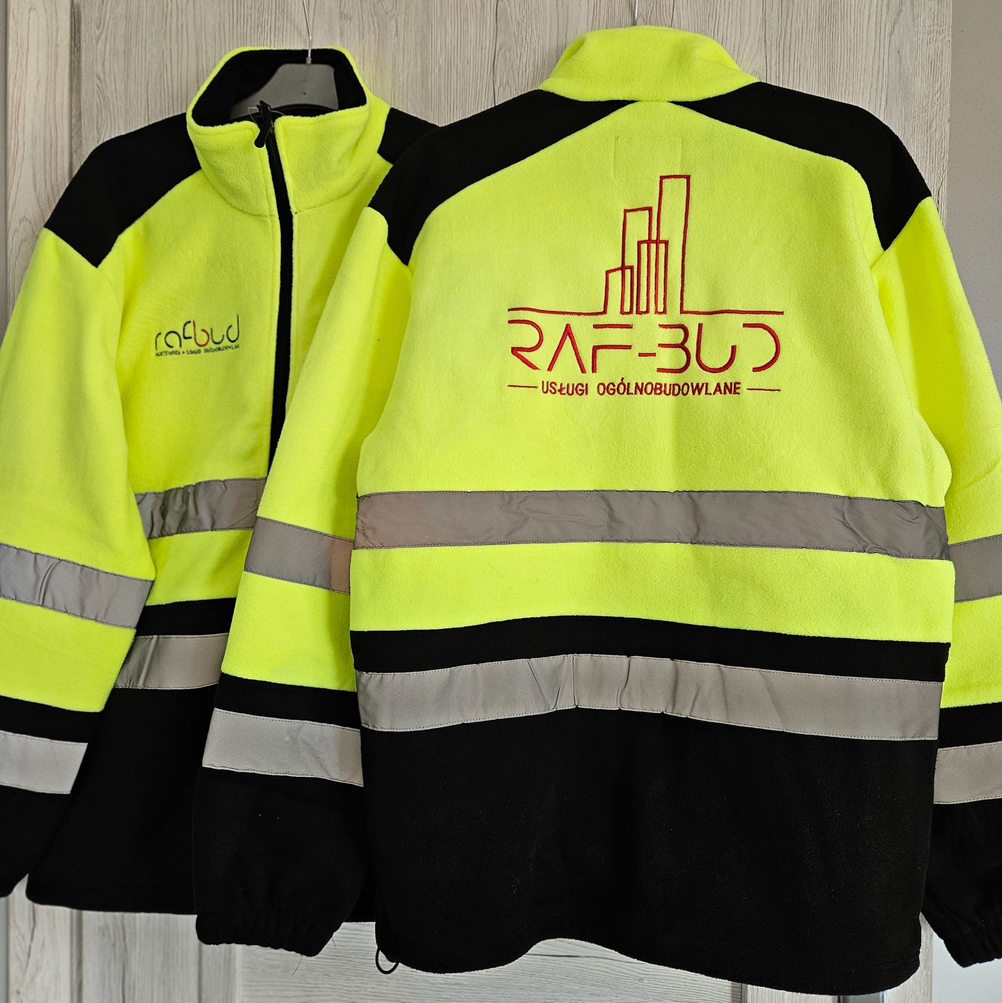 Dwie polarowe bluzy robocze w kolorze żółtym i czarnym, z odblaskowymi pasami i nadrukowanym logo firm budowlanych, wiszące na wieszakach na tle drewnianych drzwi.