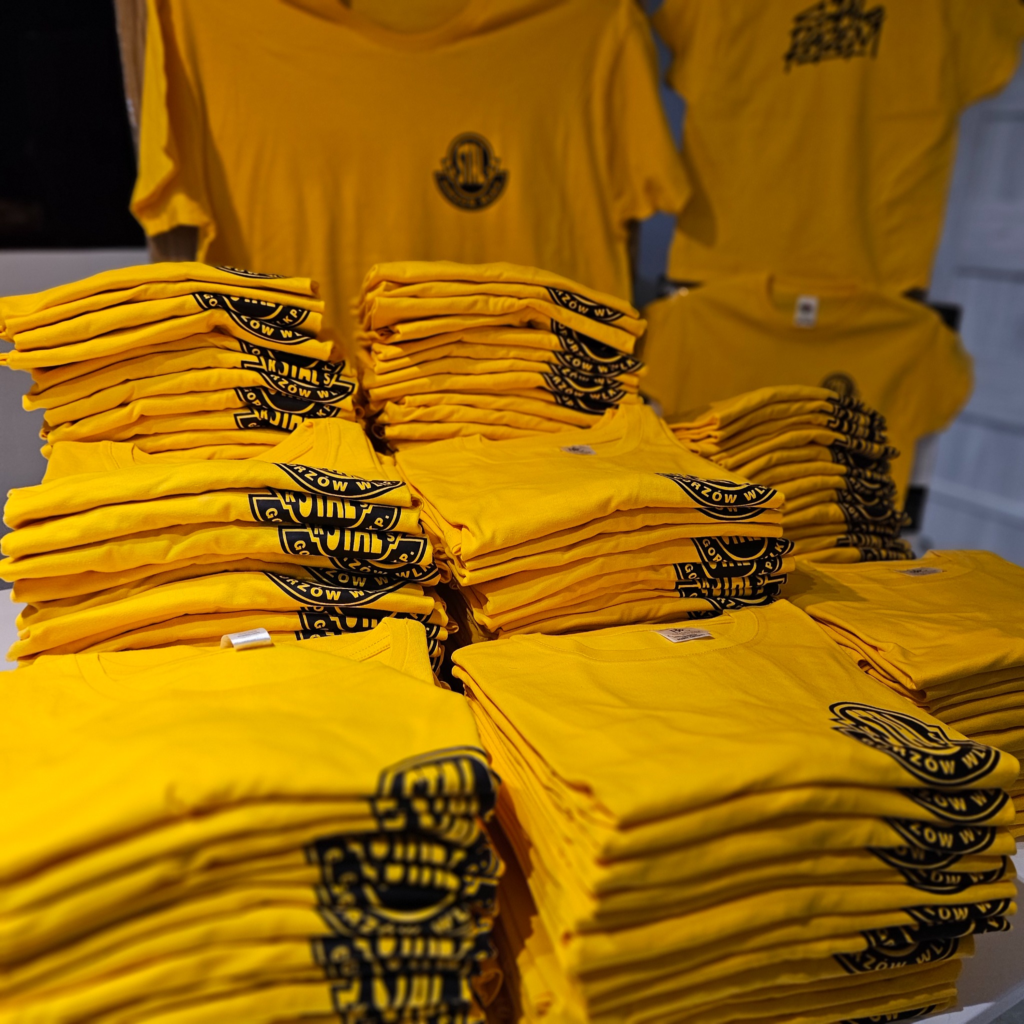 Stosy żółtych koszulek z nadrukiem w Buk. Równomiernie ułożone, gotowe do sprzedaży. Widoczne detale nadruku. U góry wiszące koszulki z logo.