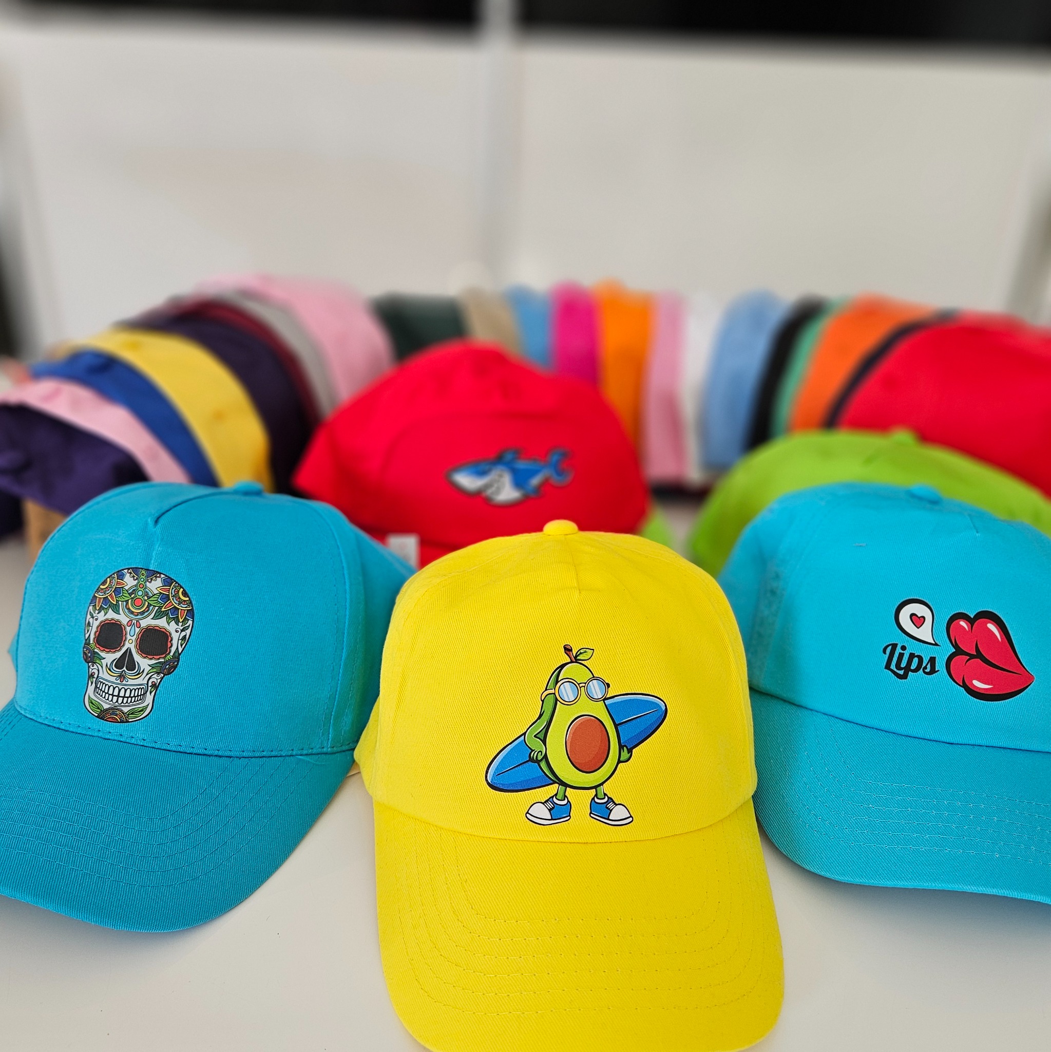 Kolorowe czapki z nadrukami: czaszka, awokado surfer, usta. W tle rząd czapek. Żółta, turkusowa i czerwona czapka na pierwszym planie. Nadruki na czapkach.