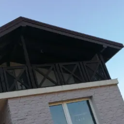 Widok z dołu na fragment domu z ciemnobrązowym dachem, drewnianą balustradą balkonu z krzyżowymi wzorami i elewacją imitującą kamień w odcieniach beżu.