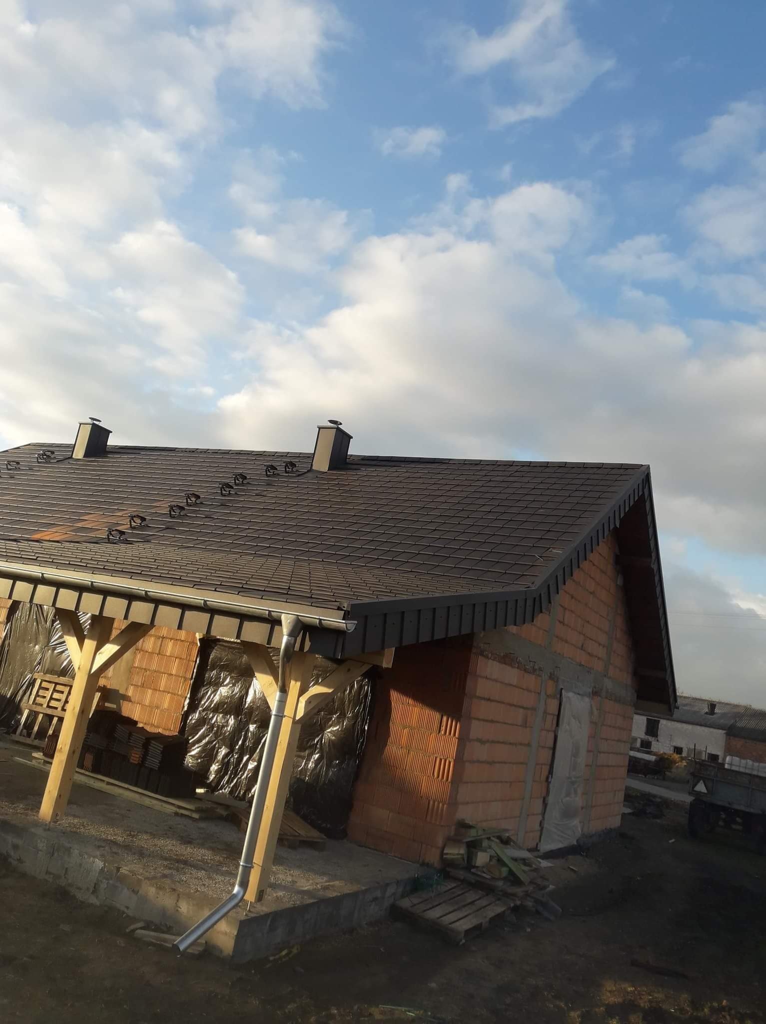 Nowy, brązowy dach z kominkami na ceglanym domu w trakcie budowy, widoczne rynny i drewniana konstrukcja pod dachem, błękitne niebo w tle.