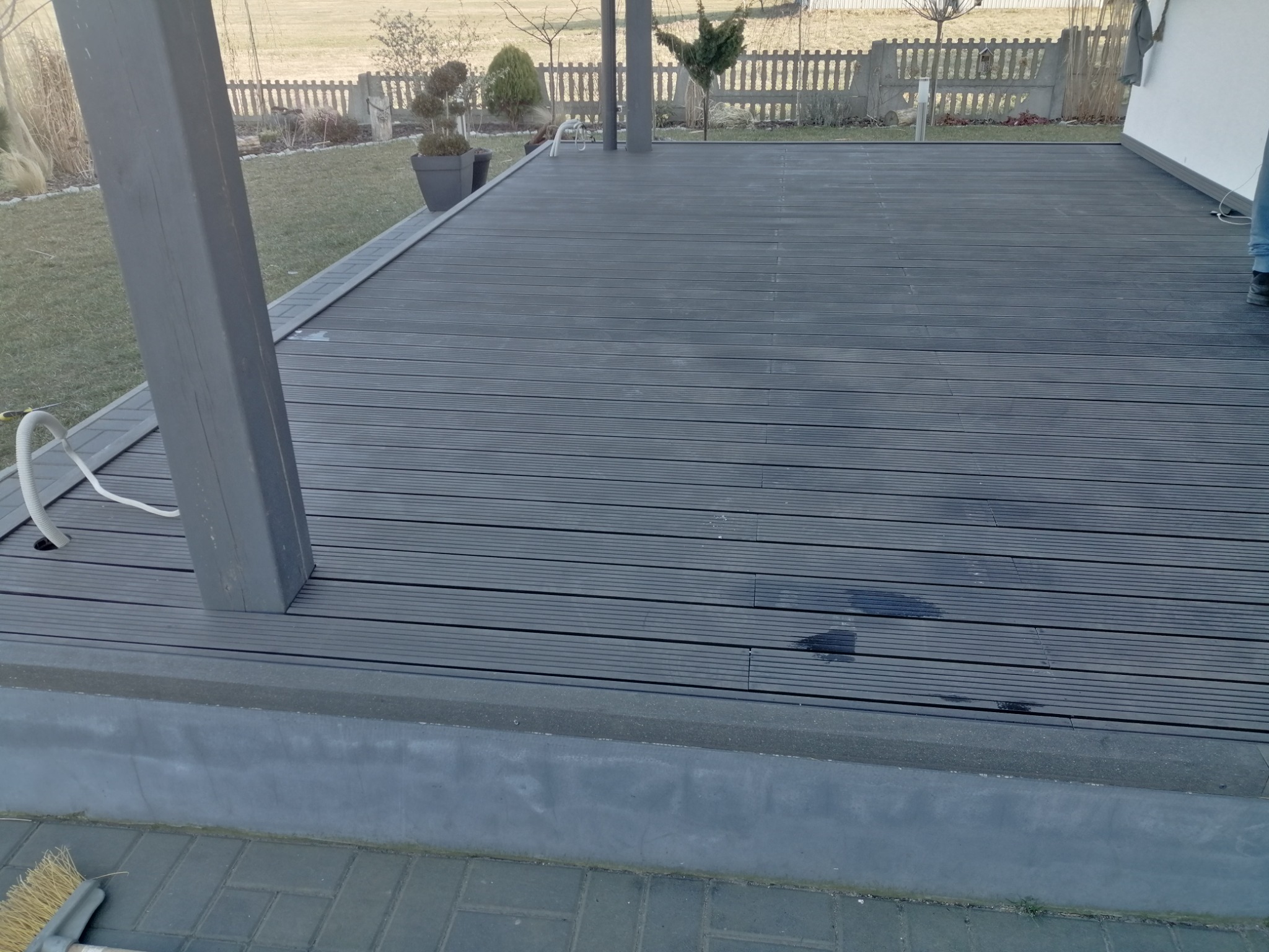 Szara, kompozytowa deska tarasowa z widocznym słupem konstrukcyjnym, obrzeżem i fragmentem elewacji domu w tle.