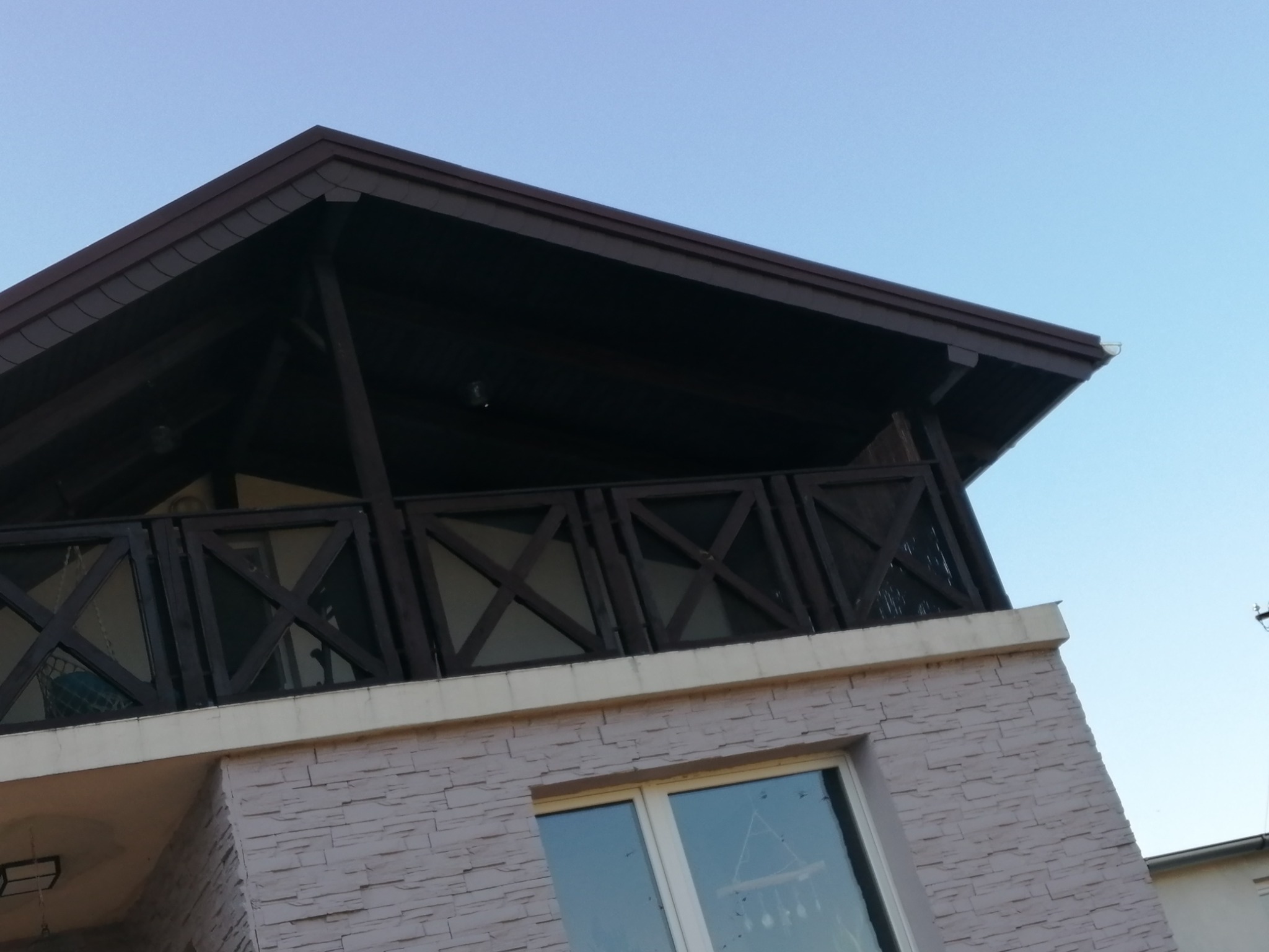 Widok z dołu na fragment domu z ciemnobrązowym dachem, drewnianą balustradą balkonu z krzyżowymi wzorami i elewacją imitującą kamień w odcieniach beżu.