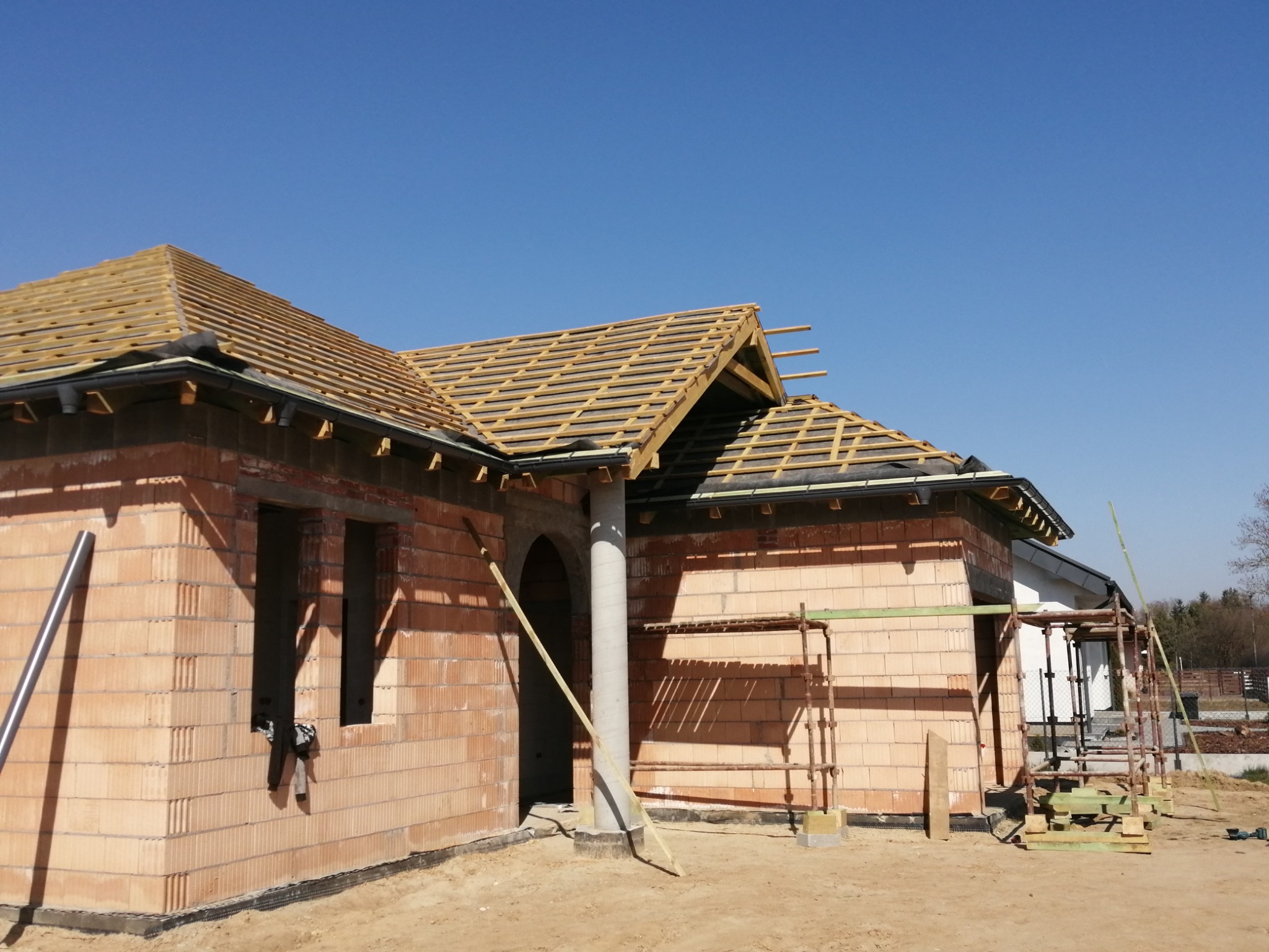 Budowa domu jednorodzinnego w trakcie, widoczne ściany z cegły ceramicznej, drewniana konstrukcja dachu z łatami, niezadaszona, rusztowania przy ścianach bocznych, błękitne niebo.