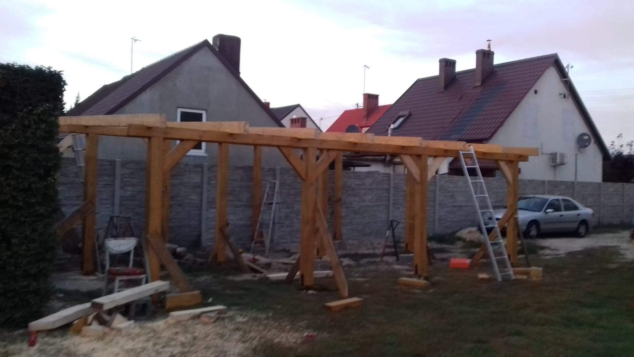 Konstrukcja drewniana z belek w trakcie budowy na tle domów jednorodzinnych i betonowego ogrodzenia, z widoczną drabiną i narzędziami.