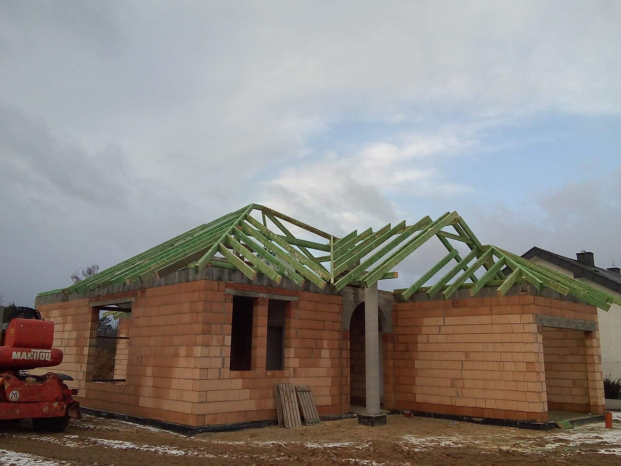 Budowa domu jednorodzinnego z czerwonej cegły, widoczna konstrukcja dachu z zielonych belek, na pierwszym planie maszyna budowlana Manitou.