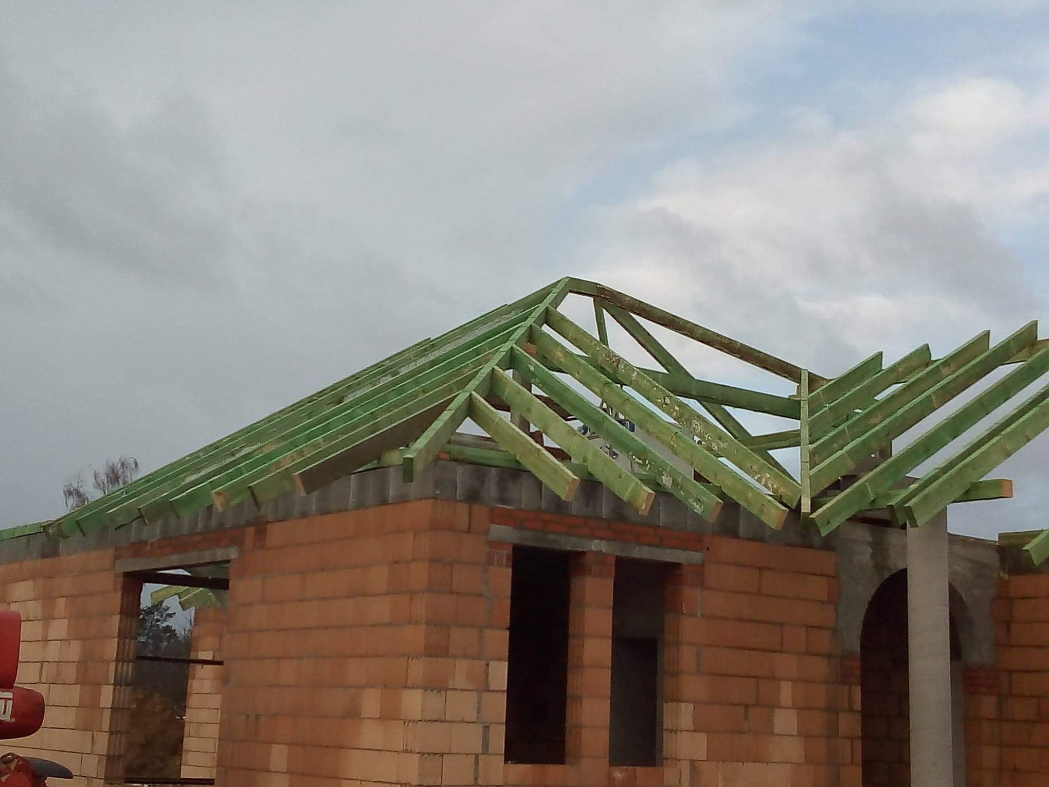 Niedokończony budynek z cegły z zieloną drewnianą konstrukcją dachu na tle pochmurnego nieba.