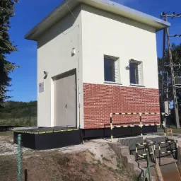 Mały budynek stacji pomp z ocieploną elewacją, pomalowaną na kremowo powyżej ceglanej części, z metalowymi drzwiami i okratowanymi oknami, stojący na betonowym fundamencie.