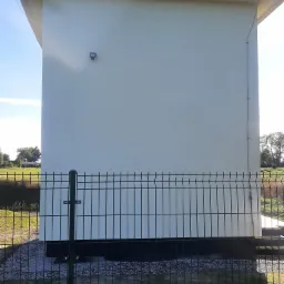 Nowo pomalowany budynek transformatorowy w kolorze kremowym, ogrodzony metalową siatką, z widocznym oświetleniem zewnętrznym i elementami instalacji elektrycznej na ścianie.