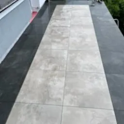 Wykończony taras z jasnoszarymi płytami na ciemnoszarym tle, widok z góry, częściowo widoczne rośliny w tle. Układ płytek wzdłuż długości tarasu.