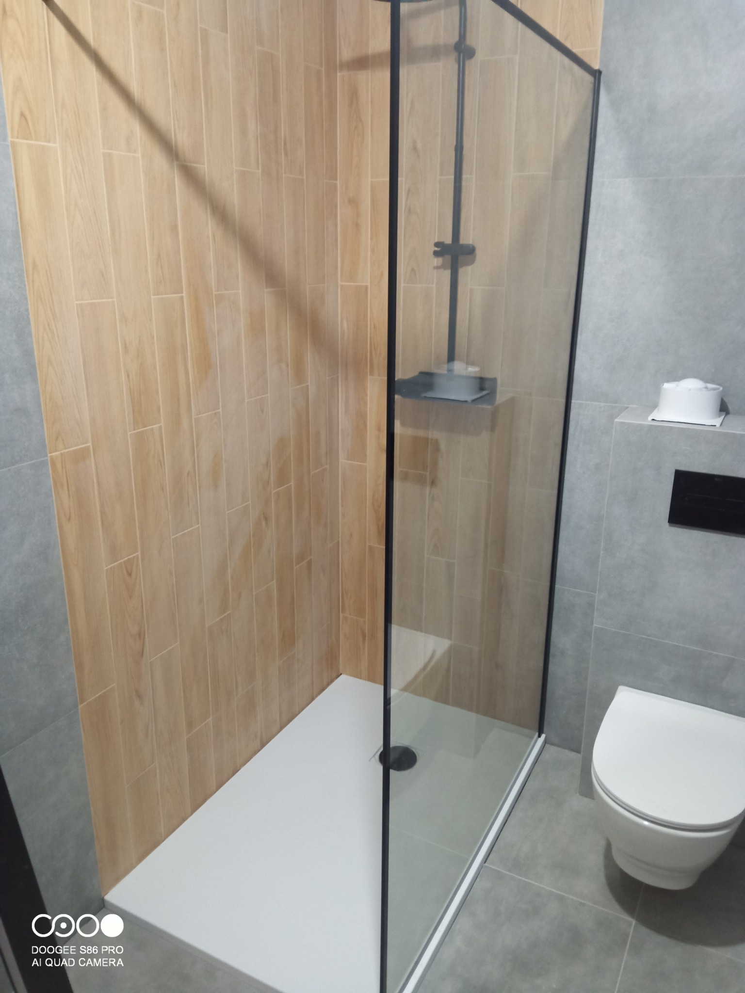 Nowoczesna kabina prysznicowa z czarnymi profilami i drewnopodobnymi płytkami na ścianie w łazience wykończonej szarymi płytkami. Widoczna toaleta.