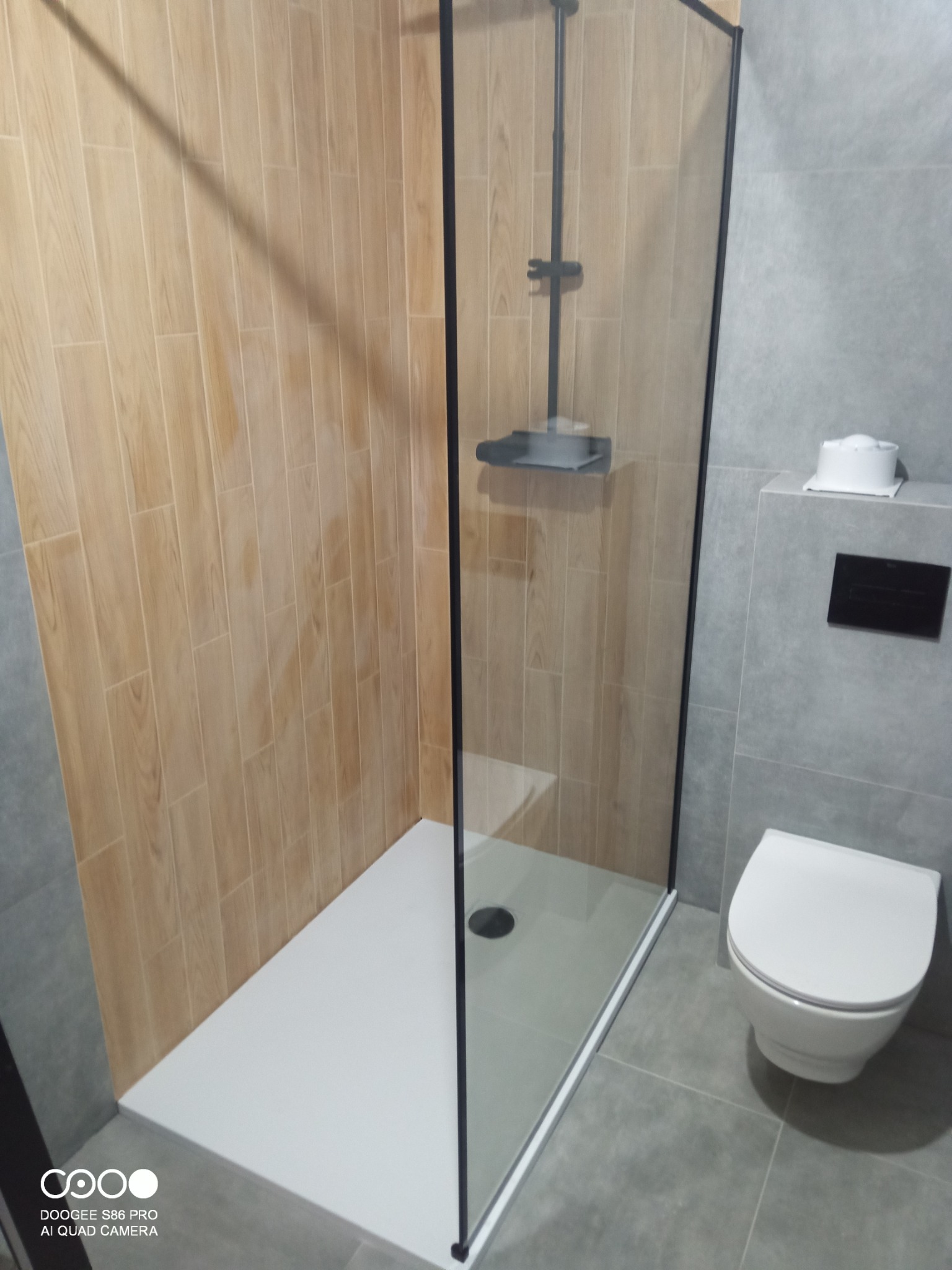 Nowoczesna kabina prysznicowa typu walk-in z czarnymi profilami, jasnymi płytkami imitującymi drewno i szarą armaturą, tworząca minimalistyczną przestrzeń łazienki.